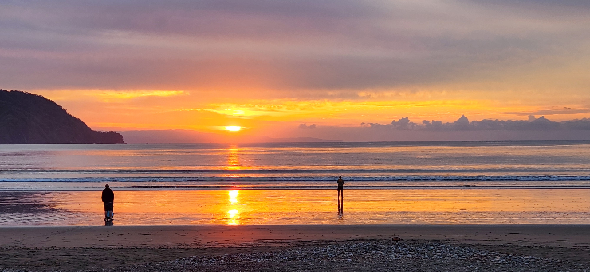 Two silhouetted figures on a beach at sunset with a vibrant orange and pink sky reflecting on the water